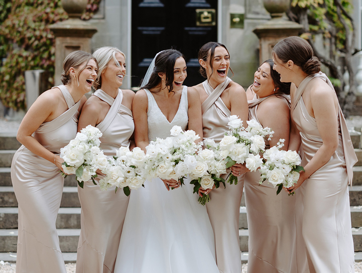 Bride and her five bridesmaids laugh together while holding white rose bouquets